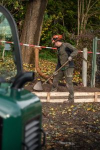 Ein Bauarbeiter in Schutzkleidung benutzt auf einer Baustelle eine Schaufel. Inmitten von Bäumen und anderen Naturelementen konzentriert er sich auf ein Baumstandortsanierungsprojekt. Rot-weißes Absperrband markiert den Hintergrund, während im Vordergrund teilweise ein grünes Baufahrzeug zu sehen ist.