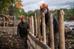 Zwei Bauarbeiter mit orangefarbenen Schutzhelmen und Gehörschutz arbeiten im Freien an einem Projekt. Im Rahmen einer Baumstandortsanierung errichten sie eine Holzbarriere aus vertikalen Baumstämmen. Die Szenerie scheint eine von Bäumen und Büschen umgebene Baustelle unter einem bewölkten Himmel zu sein.