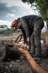Ein Arbeiter in Schutzkleidung, einschließlich Helm und Ohrenschützern, bückt sich, um auf einer schlammigen Baustelle einen dicken, hellbraunen Schlauch oder ein Rohr zu handhaben. Im Hintergrund sind Bäume und ein bewölkter Himmel zu sehen. Der Arbeiter scheint sich auf die anstehende Baumstandortsanierungsaufgabe zu konzentrieren.