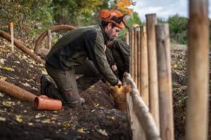Zwei Personen bauen im Freien eine Holzkonstruktion. Sie tragen Schutzhelme und Handschuhe. Der Arbeiter im Vordergrund befestigt ein seilartiges Material an der Konstruktion. Die Szene, die an ein Baumstandortsanierungsprojekt erinnert, spielt sich in einer natürlichen Umgebung ab, in der grünes Laub und Herbstblätter verstreut sind.