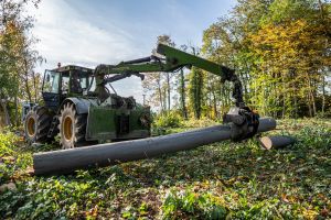 Ein grüner Traktor mit einem Holzgreifer hebt an einem sonnigen Tag zwei große Baumstämme auf einer Waldlichtung an. Dies ist Teil der laufenden Baumfällung. Der Boden ist mit Blättern und Zweigen bedeckt und die umliegenden Bäume haben grünes und gelbes Laub.