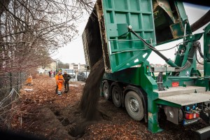 Ein großer grüner Muldenkipper lädt einen Haufen Erde auf einen Weg ab, wahrscheinlich um den Bereich für eine neue Bepflanzung vorzubereiten. In der Nähe sind mehrere Arbeiter in orangefarbener Schutzausrüstung zu sehen, einige stehen näher am Laster und andere arbeiten weiter unten am Weg. Der Bereich ist von kahlen Bäumen und einem Metallzaun begrenzt.