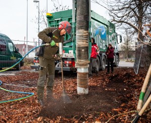 Ein Arbeiter mit Gehörschutz bedient ein Bodeninjektionsgerät neben einem Baum, dessen Stamm mit einem Schutz umwickelt ist, um die Bodenverbesserung zu verbessern. In der Nähe steht ein grüner Lastwagen mit der Aufschrift „BLANKINGBA“, und zwei Beobachter stehen da und beobachten ihn. Der Boden ist mit braunen Blättern und Mulch bedeckt.