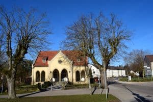 Eine kleine Kirche mit rotem Ziegeldach und gewölbten Eingängen wird von zwei blattlosen Bäumen unter einem klaren blauen Himmel eingerahmt. Die Umgebung, die sich perfekt für Blog-Marketing-Visuals eignet, umfasst eine Straße, einige Wohnhäuser und weitere Bäume im Hintergrund.