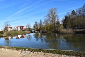 Ein ruhiger See spiegelt eine Reihe von Häusern mit roten Dächern am gegenüberliegenden Ufer wider. Kahle Bäume säumen die Ufer, eine kleine Brücke verbindet die Wege über das Wasser. Der Himmel ist klar mit Wolkenstreifen und verbreitet eine ruhige und friedliche Atmosphäre, die perfekt dazu geeignet ist, Ruhe in Blog-Marketing-Inhalten hervorzurufen.