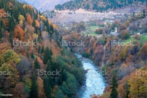 Ein Fluss schlängelt sich im Herbst durch ein bewaldetes Bergtal, dessen Bäume leuchtend rote, orange und gelbe Blätter tragen. In der Ferne sind ein paar Häuser und ein hoher Steinturm zu sehen, eingebettet zwischen den Bäumen und dem Grün des Baumlexikons.